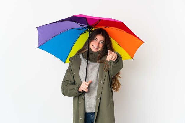 Jovem mulher caucasiana segurando um guarda-chuva isolado na parede branca e apontando para a frente com uma expressão feliz