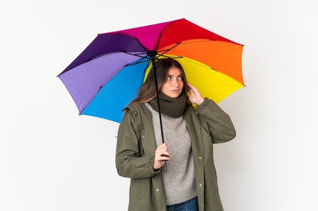 Jovem mulher caucasiana segurando um guarda-chuva isolado na parede branca, tendo dúvidas