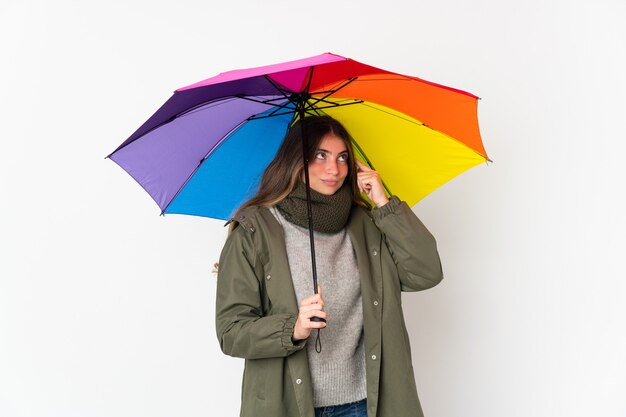 Jovem mulher caucasiana segurando um guarda-chuva isolado no fundo branco, tendo dúvidas e pensando