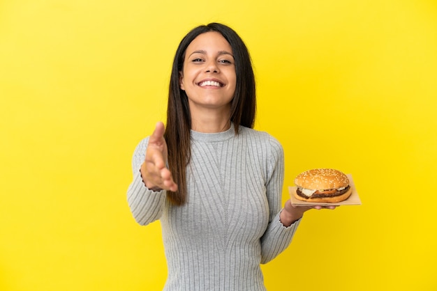 Jovem mulher caucasiana segurando um hambúrguer isolado em um fundo amarelo, apertando as mãos para fechar um bom negócio