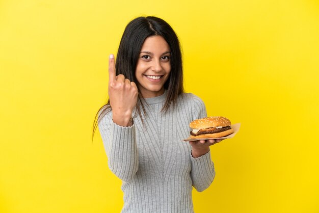 Jovem mulher caucasiana segurando um hambúrguer isolado em um fundo amarelo, fazendo um gesto de aproximação