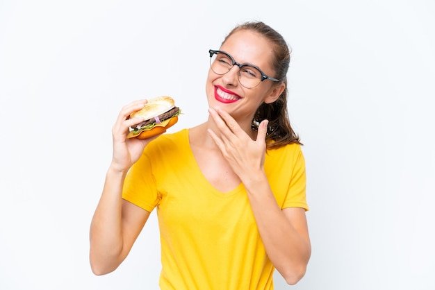 Jovem mulher caucasiana segurando um hambúrguer isolado no fundo branco olhando para cima enquanto sorria