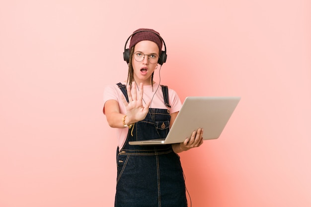 Jovem mulher caucasiana, segurando um laptop em pé com a mão estendida, mostrando o sinal de stop, impedindo-o.