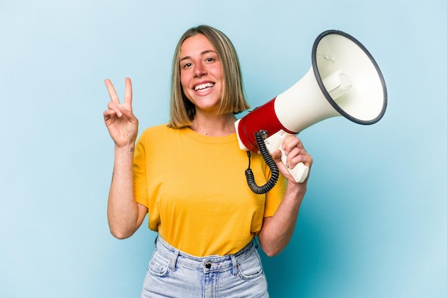 Jovem mulher caucasiana segurando um megafone isolado em fundo azul alegre e despreocupado, mostrando um símbolo de paz com os dedos