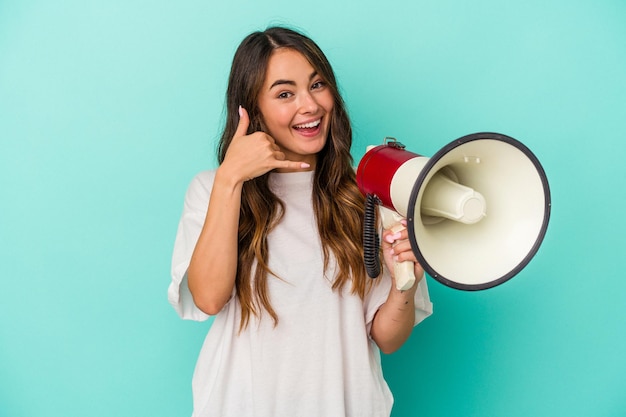 Jovem mulher caucasiana segurando um megafone isolado em fundo azul, mostrando um gesto de chamada de celular com os dedos.