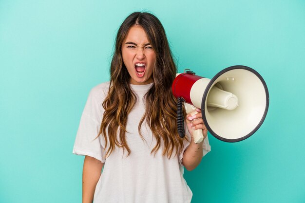 Jovem mulher caucasiana segurando um megafone isolado no fundo azul, gritando muito irritado e agressivo.