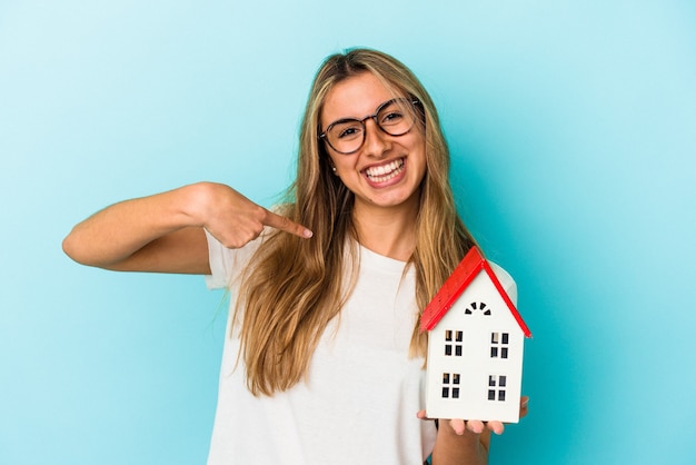 Jovem mulher caucasiana segurando um modelo de casa isolada na parede azul pessoa apontando com a mão para um espaço de cópia de camisa, orgulhosa e confiante