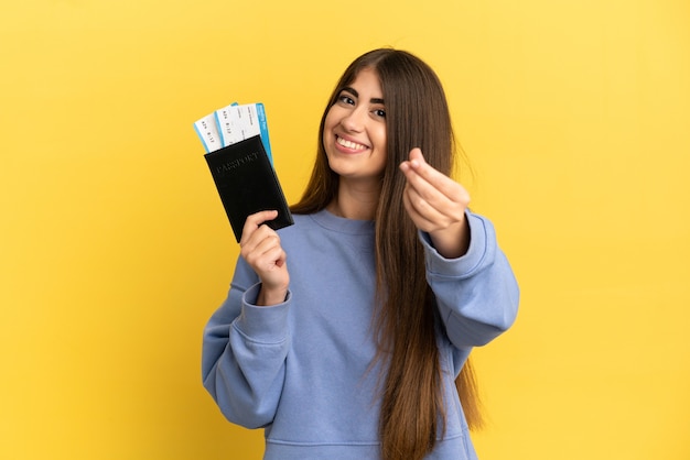 Jovem mulher caucasiana segurando um passaporte isolado em um fundo amarelo fazendo gesto de dinheiro