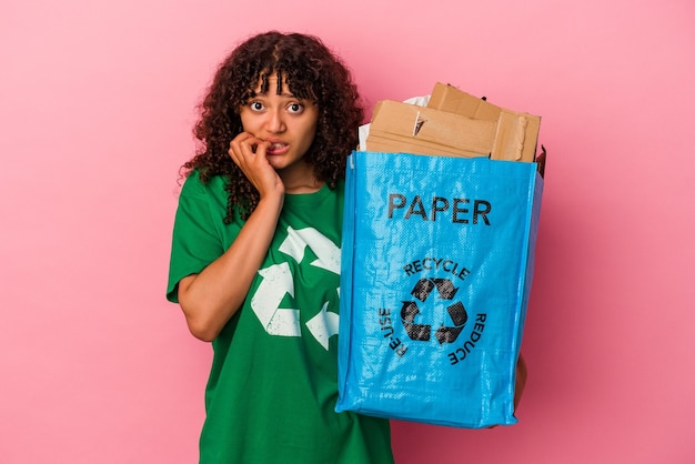 Jovem mulher caucasiana segurando um plástico reciclado isolado na parede rosa, roendo as unhas, nervosa e muito ansiosa.