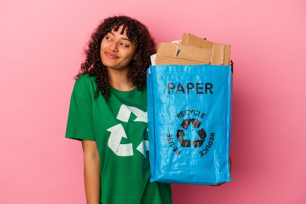 Jovem mulher caucasiana segurando um plástico reciclado isolado na parede rosa, sonhando em alcançar objetivos e propósitos