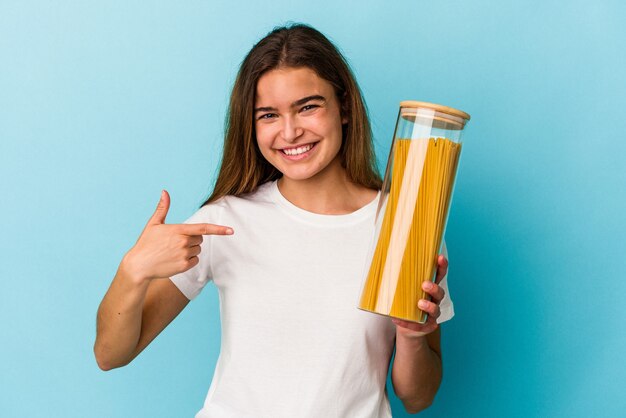 Foto jovem mulher caucasiana segurando um pote de macarrão isolado em um fundo azul pessoa apontando com a mão para um espaço de cópia de camisa, orgulhosa e confiante