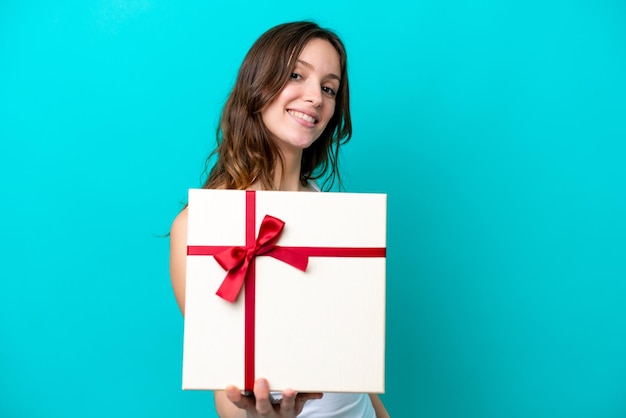 Jovem mulher caucasiana segurando um presente isolado em fundo azul com expressão feliz