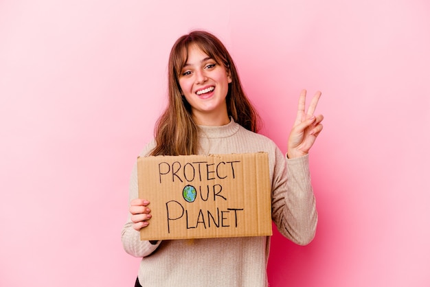 Jovem mulher caucasiana segurando um Proteger nosso cartaz de planeta isolado alegre e despreocupado, mostrando um símbolo de paz com os dedos.