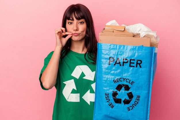 Jovem mulher caucasiana segurando um saco de papelão reciclado isolado no fundo rosa com os dedos nos lábios, mantendo um segredo.