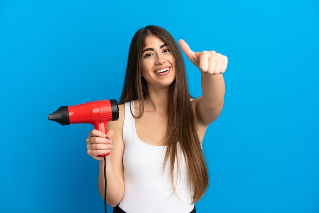 Jovem mulher caucasiana segurando um secador de cabelo isolado em um fundo azul com o polegar para cima porque algo de bom aconteceu