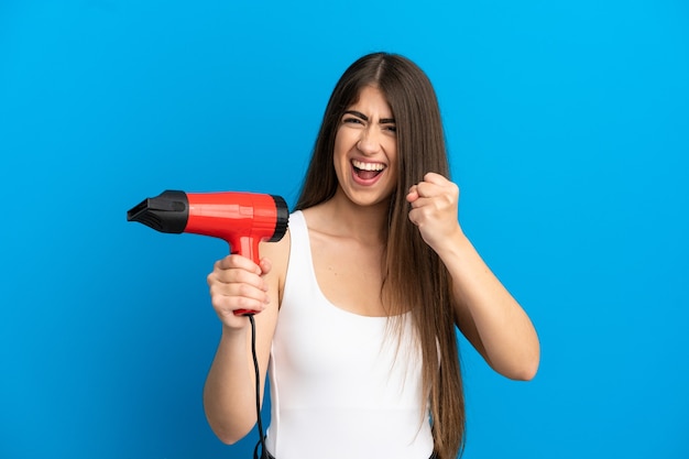 Jovem mulher caucasiana segurando um secador de cabelo isolado em um fundo azul, comemorando uma vitória