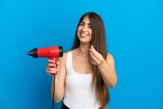 Jovem mulher caucasiana segurando um secador de cabelo isolado em um fundo azul, fazendo gesto de dinheiro