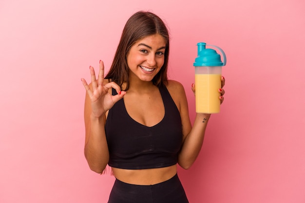 Jovem mulher caucasiana segurando um shake de proteína isolado na parede amarela alegre e confiante, mostrando um gesto de ok.