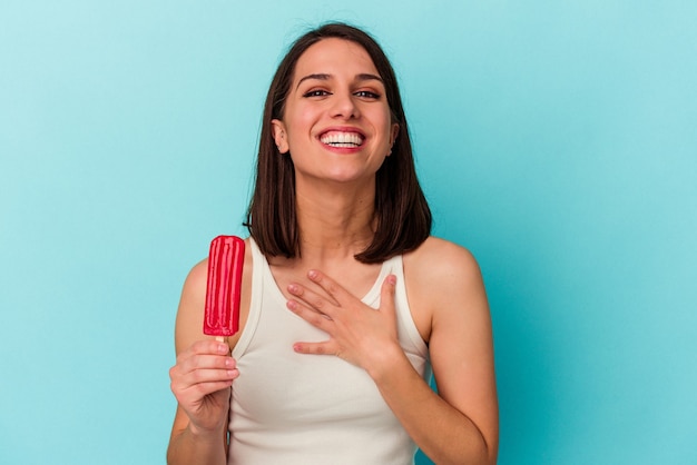 Jovem mulher caucasiana segurando um sorvete isolado no fundo azul ri alto, mantendo a mão no peito.
