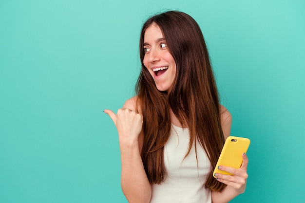 Jovem mulher caucasiana segurando um telefone celular isolado em pontos de fundo azul com o dedo polegar afastado, rindo e despreocupada.