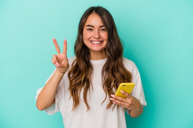 Jovem mulher caucasiana segurando um telefone celular isolado em um fundo azul, mostrando o número dois com os dedos.