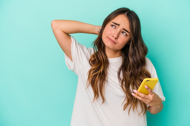 Jovem mulher caucasiana, segurando um telefone celular isolado em um fundo azul, tocando a parte de trás da cabeça, pensando e fazendo uma escolha.