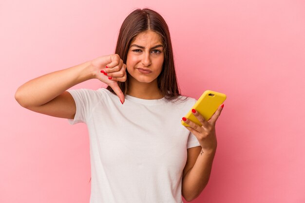 Jovem mulher caucasiana segurando um telefone celular isolado em um fundo rosa, mostrando um gesto de antipatia, polegares para baixo. Conceito de desacordo.