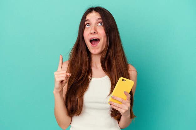 Foto jovem mulher caucasiana, segurando um telefone celular isolado na parede azul apontando para cima com a boca aberta.