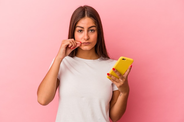 Jovem mulher caucasiana, segurando um telefone celular isolado na parede rosa com os dedos nos lábios, mantendo um segredo.