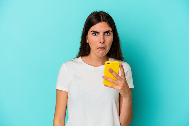 Jovem mulher caucasiana segurando um telefone celular isolado no fundo azul encolhe os ombros e abre os olhos confusos.