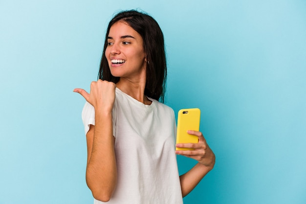 Jovem mulher caucasiana segurando um telefone móvel isolado em pontos de fundo azul com o dedo polegar afastado, rindo e despreocupada.