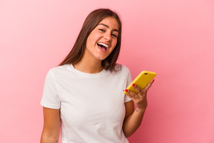 Foto jovem mulher caucasiana, segurando um telefone móvel isolado no fundo rosa, rindo e se divertindo.