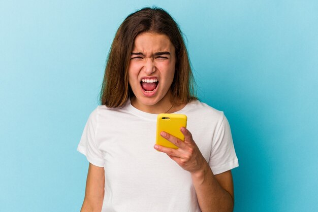 Jovem mulher caucasiana segurando um telefone móvel isolado sobre fundo azul, gritando muito zangada e agressiva.