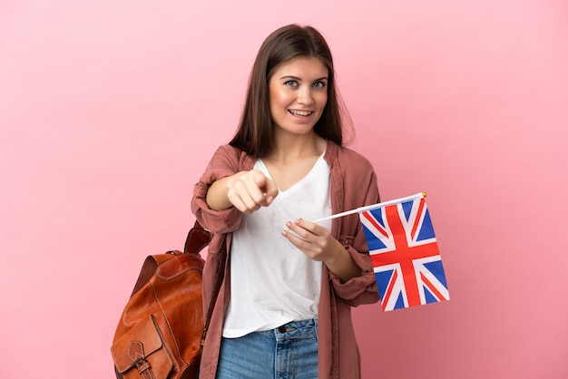 Jovem mulher caucasiana segurando uma bandeira do Reino Unido isolada em um fundo rosa apontando para a frente com uma expressão feliz