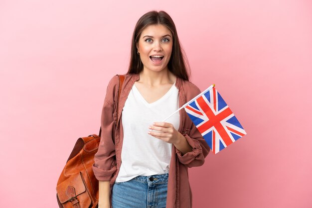 Jovem mulher caucasiana segurando uma bandeira do Reino Unido isolada em um fundo rosa com expressão facial de surpresa