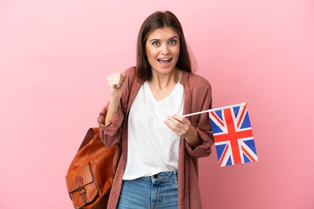 Jovem mulher caucasiana segurando uma bandeira do Reino Unido isolada em um fundo rosa, comemorando a vitória na posição de vencedora