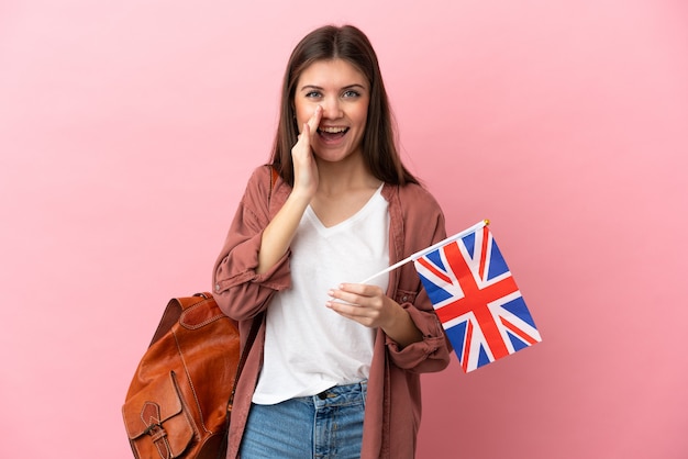Jovem mulher caucasiana segurando uma bandeira do Reino Unido isolada em um fundo rosa gritando com a boca bem aberta