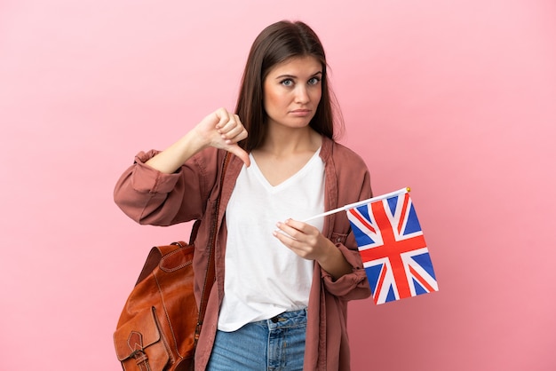 Jovem mulher caucasiana segurando uma bandeira do reino unido isolada em um fundo rosa, mostrando o polegar para baixo com expressão negativa