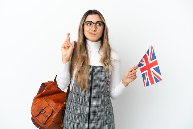 Jovem mulher caucasiana segurando uma bandeira do Reino Unido, isolada no fundo branco, cruzando os dedos e desejando o melhor