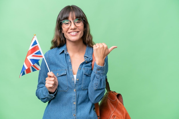 Jovem mulher caucasiana segurando uma bandeira do Reino Unido sobre fundo isolado, apontando para o lado para apresentar um produto