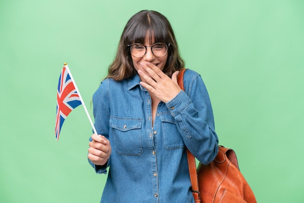 Jovem mulher caucasiana, segurando uma bandeira do Reino Unido sobre fundo isolado, feliz e sorridente, cobrindo a boca com a mão