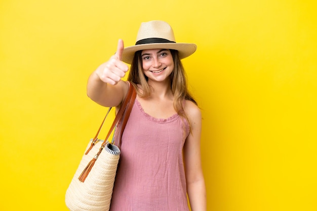 Jovem mulher caucasiana segurando uma bolsa de praia isolada em fundo amarelo com polegares para cima porque algo bom aconteceu