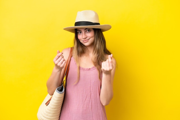 Foto jovem mulher caucasiana, segurando uma bolsa de praia isolada em fundo amarelo, fazendo gesto de dinheiro