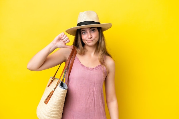 Jovem mulher caucasiana segurando uma bolsa de praia isolada em fundo amarelo, mostrando o polegar para baixo com expressão negativa