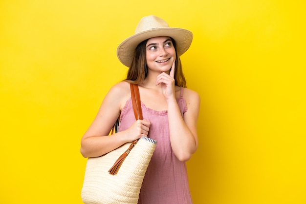 Jovem mulher caucasiana segurando uma bolsa de praia isolada em fundo amarelo pensando em uma ideia enquanto olha para cima