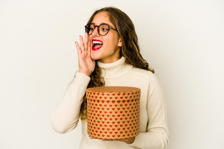 Foto jovem mulher caucasiana segurando uma caixa de dia dos namorados coração isolada gritando e segurando a palma da mão perto da boca aberta.
