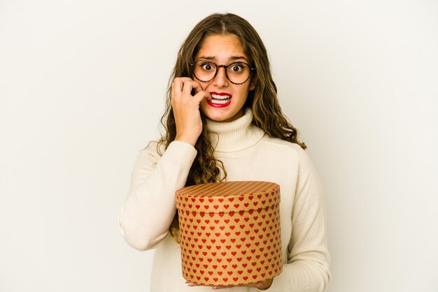 Jovem mulher caucasiana, segurando uma caixa de dia dos namorados coração isolada unhas roendo, nervosa e muito ansiosa.