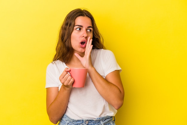 Jovem mulher caucasiana, segurando uma caneca isolada em um fundo amarelo, está contando uma notícia secreta da frenagem e olhando para o lado