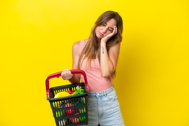 Foto jovem mulher caucasiana segurando uma cesta de compras cheia de comida isolada em fundo amarelo com dor de cabeça
