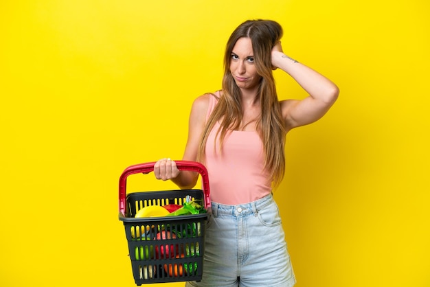 Foto jovem mulher caucasiana segurando uma cesta de compras cheia de comida isolada em fundo amarelo com dúvidas